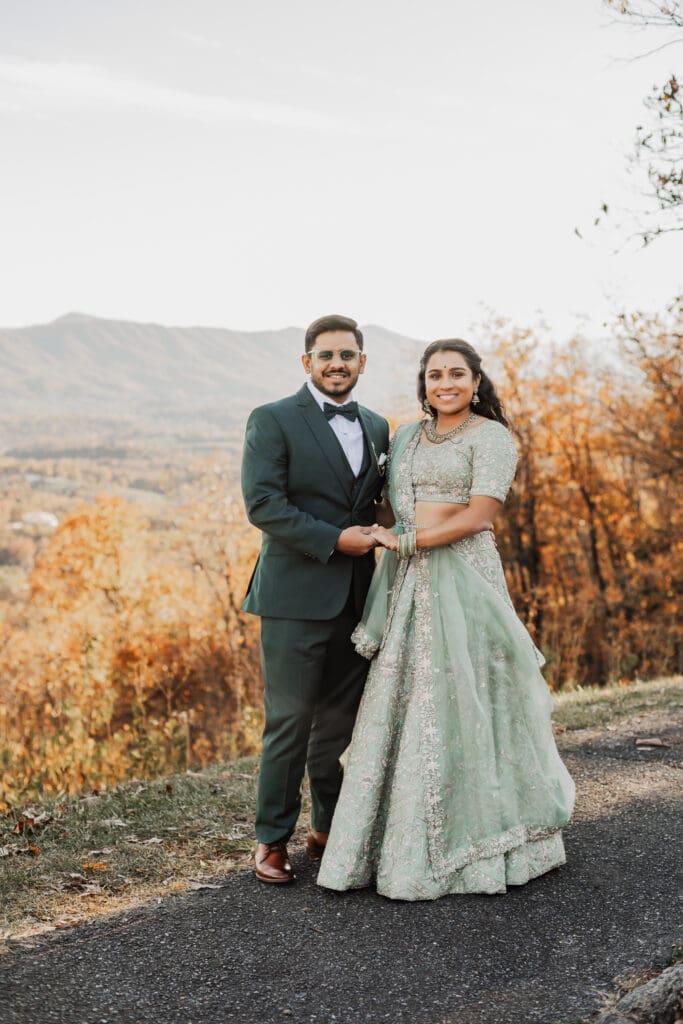 Indian couple in a in green wedding tux and lehenga in a field of green grass with a rust colored mountain in the background with blue rolling mountains in the background. They are standing close and kissing.