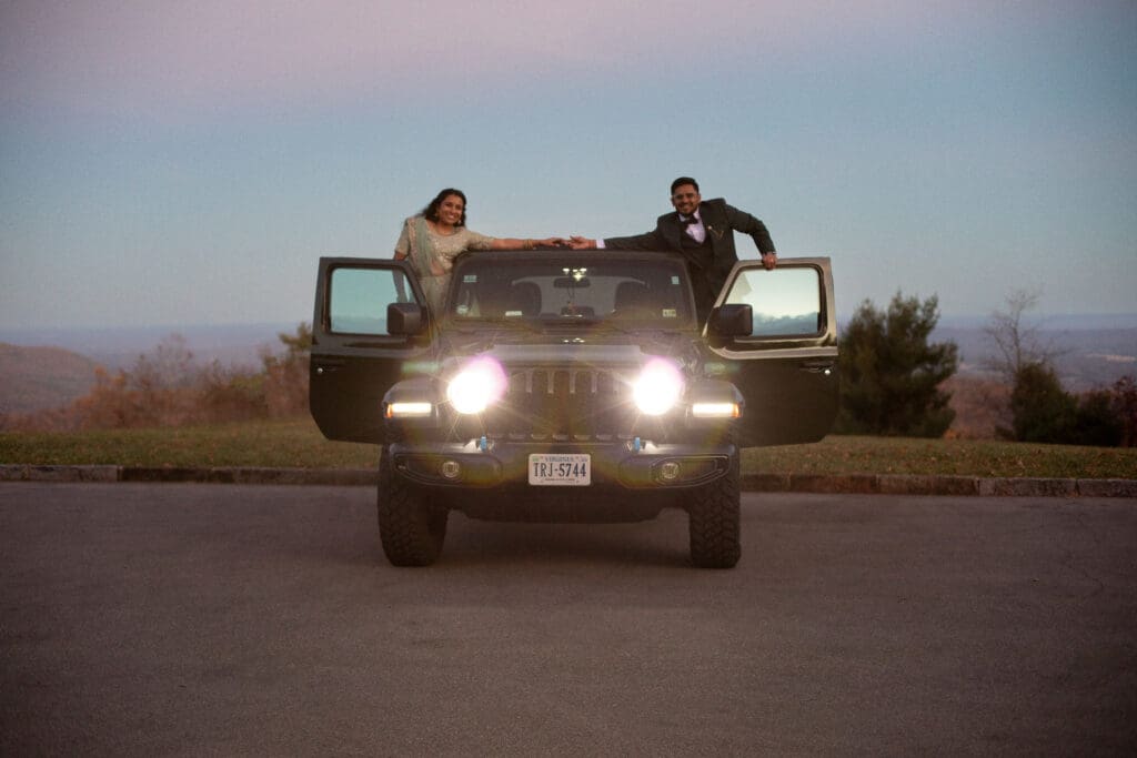 Indian couple in a in green wedding tux and lehenga in a field of green grass with a rust colored mountain in the background with blue rolling mountains in the background. They are in the dark being illuminated by the jeep lights hanging out the doors and holding hands over the roof