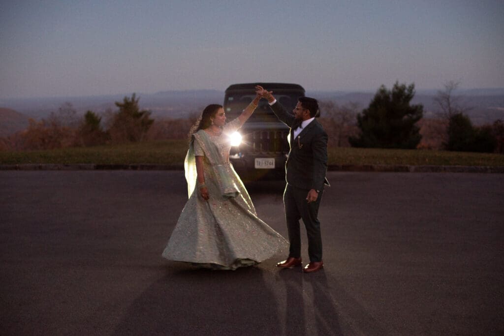 Indian couple in a in green wedding tux and lehenga in a field of green grass with a rust colored mountain in the background with blue rolling mountains in the background. They are in the dark being illuminated by the jeep lights and dancing.
