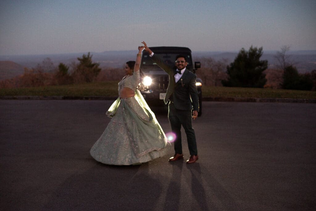 Indian couple in a in green wedding tux and lehenga in a field of green grass with a rust colored mountain in the background with blue rolling mountains in the background. They are in the dark being illuminated by the jeep lights and dancing.