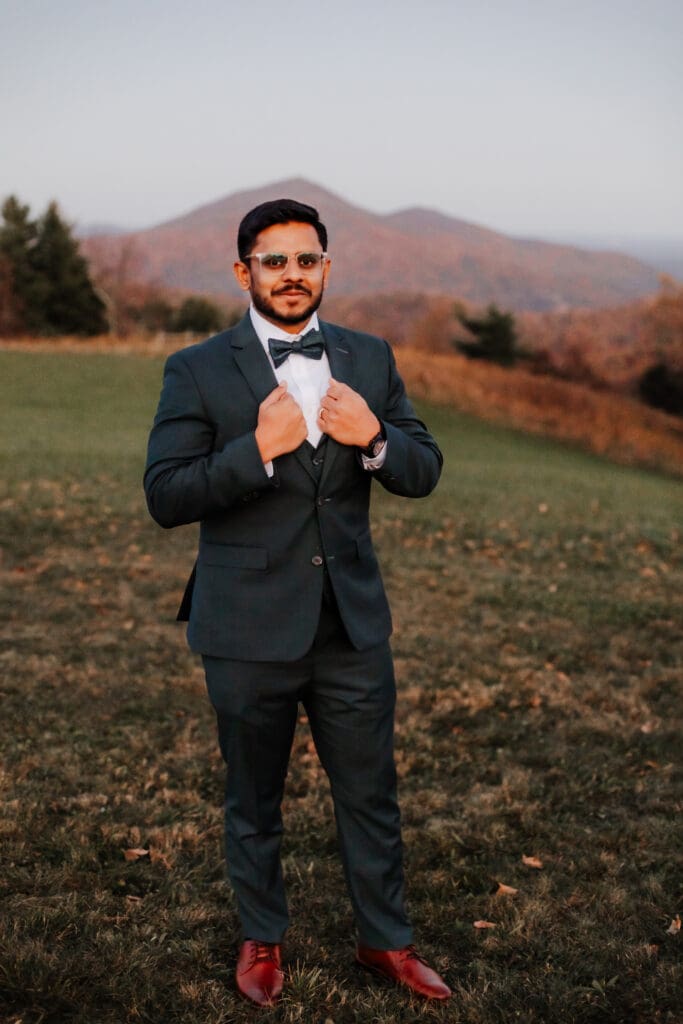 Indian groom in a in green wedding tux in a field of green grass with a rust colored mountain in the background with blue rolling mountains in the background.