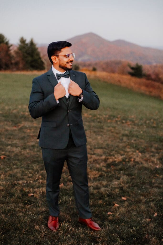 Indian groom in a in green wedding tux in a field of green grass with a rust colored mountain in the background with blue rolling mountains in the background.