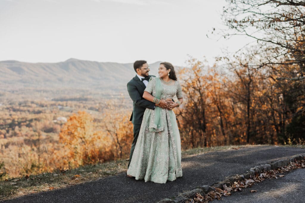 Indian couple in a in green wedding tux and lehenga in a field of green grass with a rust colored mountain in the background with blue rolling mountains in the background. They are standing close and kissing.