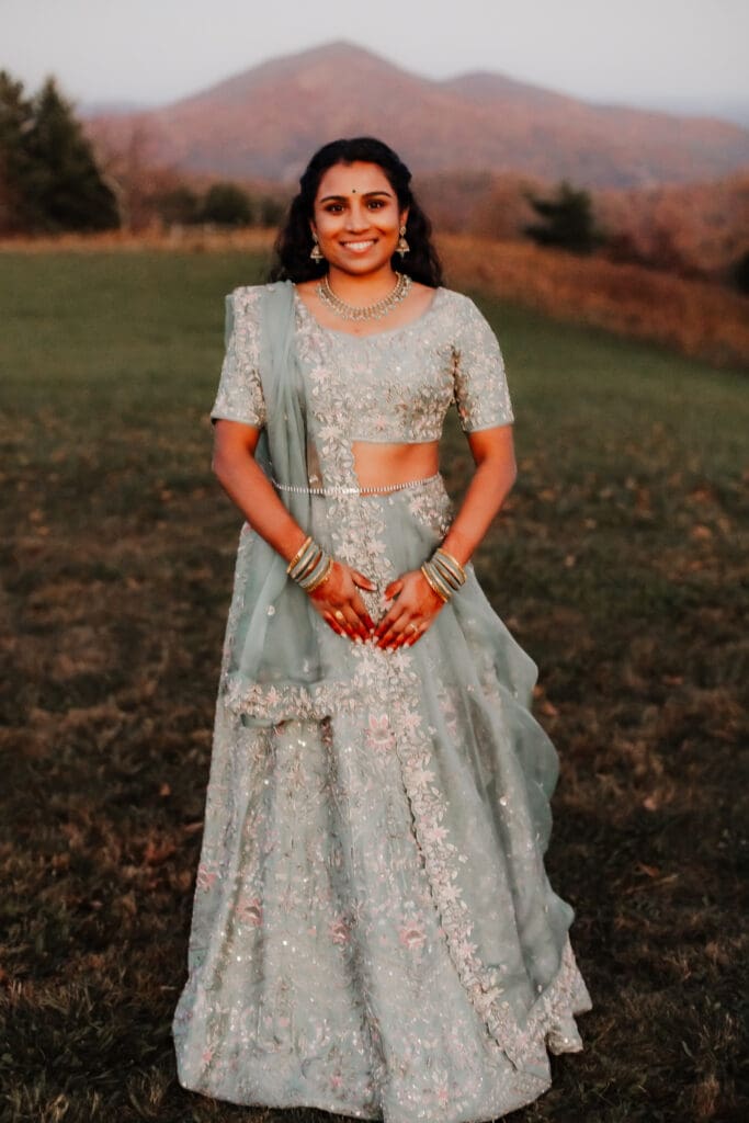 Indian bride in a Lehenga in green wedding attire in a field of green grass with a rust colored mountain in the background with blue rolling mountains in the background.