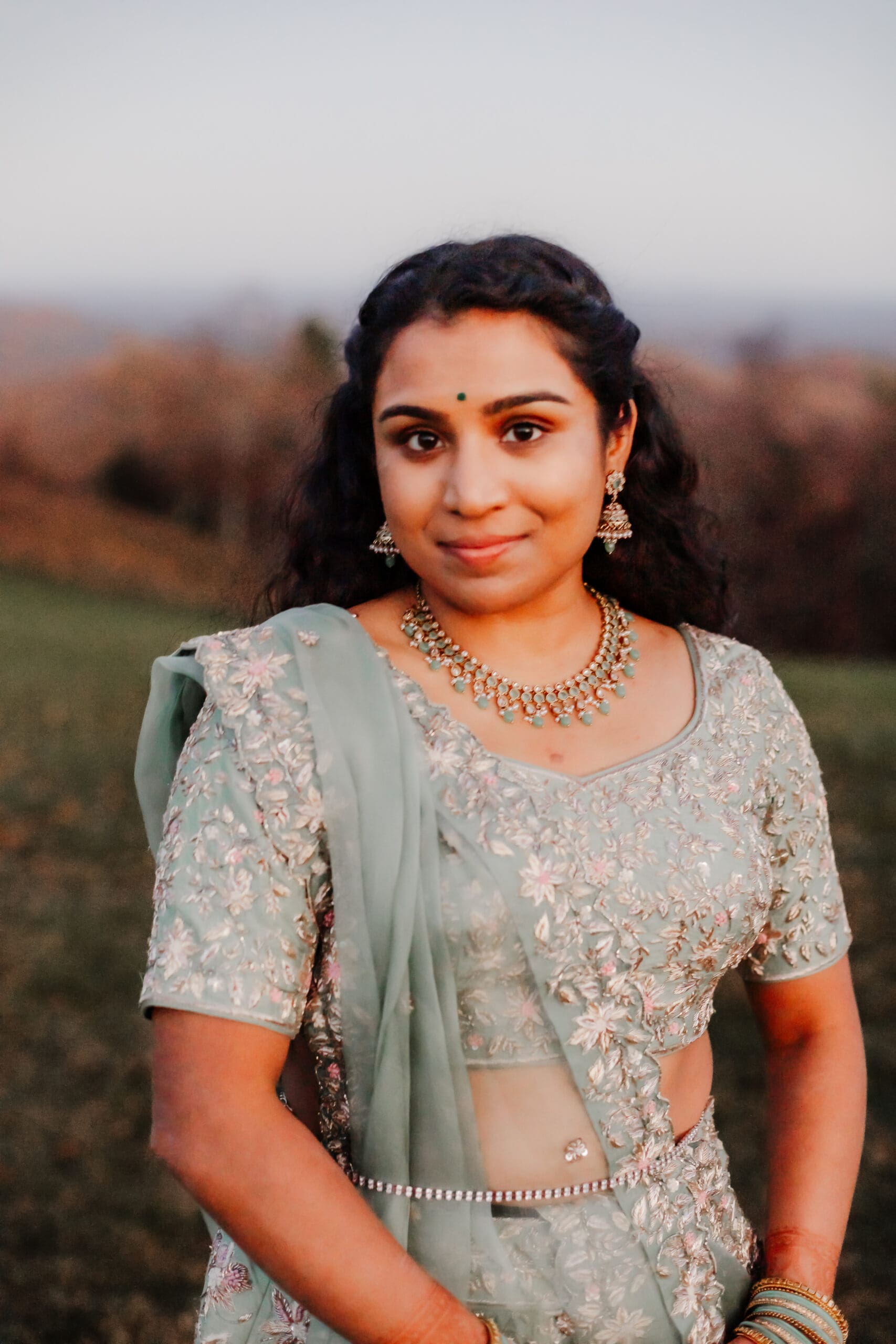 Indian bride in a Lehenga in green wedding attire in a field of green grass with a rust colored mountain in the background with blue rolling mountains in the background.
