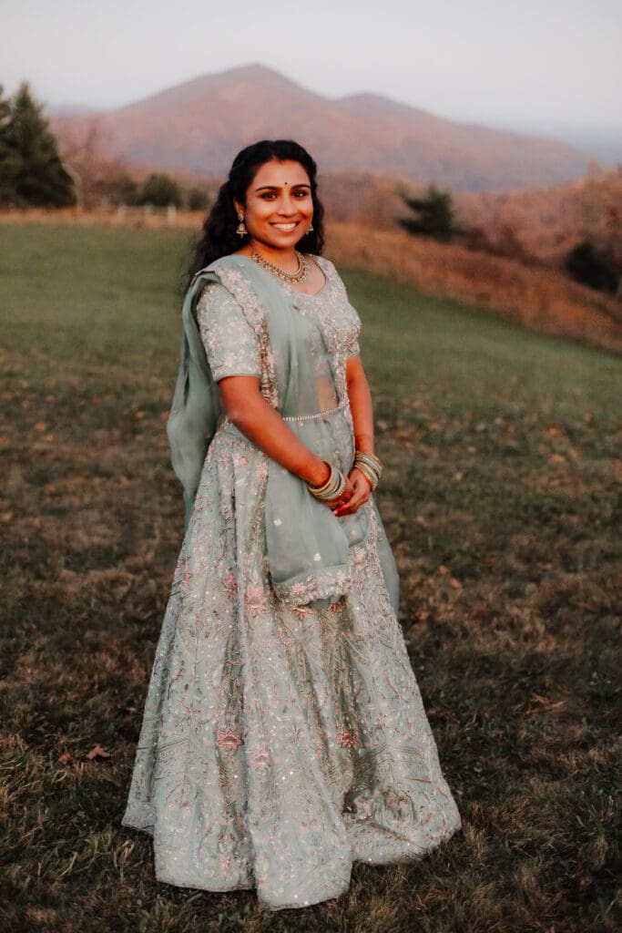 Indian bride in a Lehenga in green wedding attire in a field of green grass with a rust colored mountain in the background with blue rolling mountains in the background.