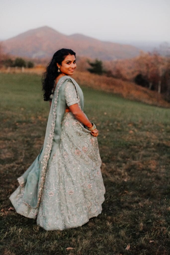Indian bride in a Lehenga doing the Sangeet dance in green wedding attire in a field of green grass with a rust colored mountain in the background with blue rolling mountains in the background.