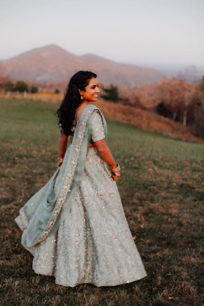 Indian bride in a Lehenga doing the Sangeet dance in green wedding attire in a field of green grass with a rust colored mountain in the background with blue rolling mountains in the background.