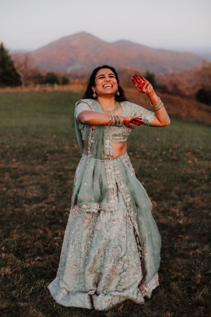 Indian bride in a Lehenga doing the Sangeet dance in green wedding attire in a field of green grass with a rust colored mountain in the background with blue rolling mountains in the background.