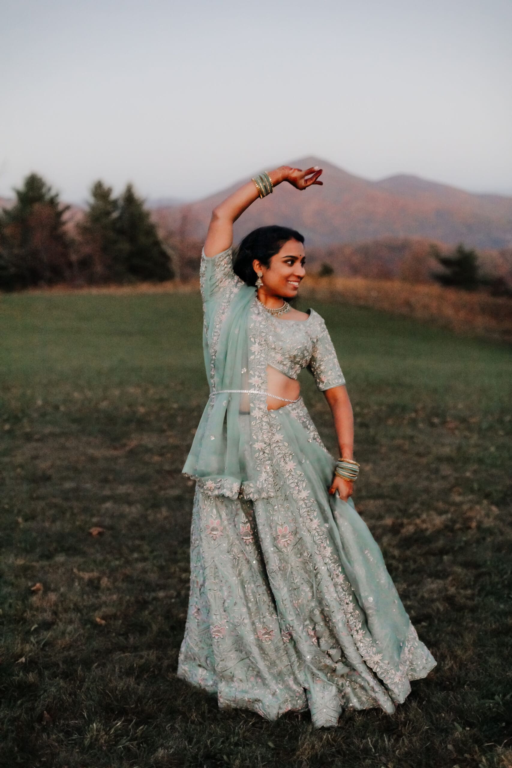 Indian bride in a Lehenga doing the Sangeet dance in green wedding attire in a field of green grass with a rust colored mountain in the background with blue rolling mountains in the background.