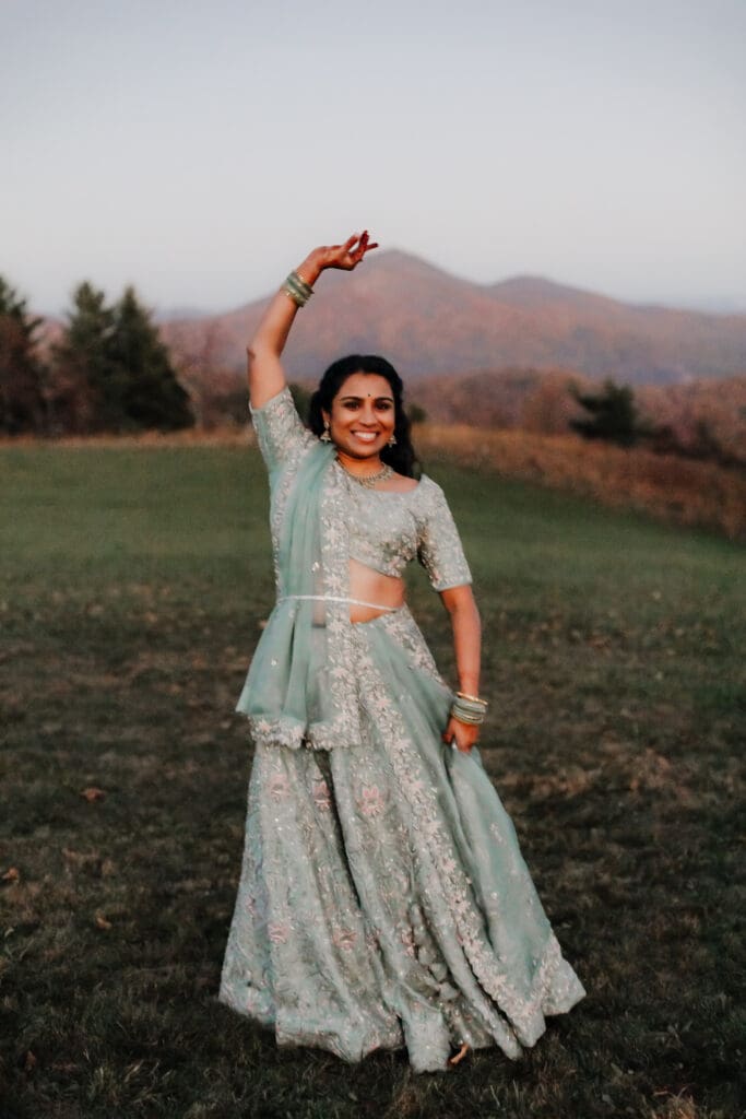 Indian bride in a Lehenga doing the Sangeet dance in green wedding attire in a field of green grass with a rust colored mountain in the background with blue rolling mountains in the background.