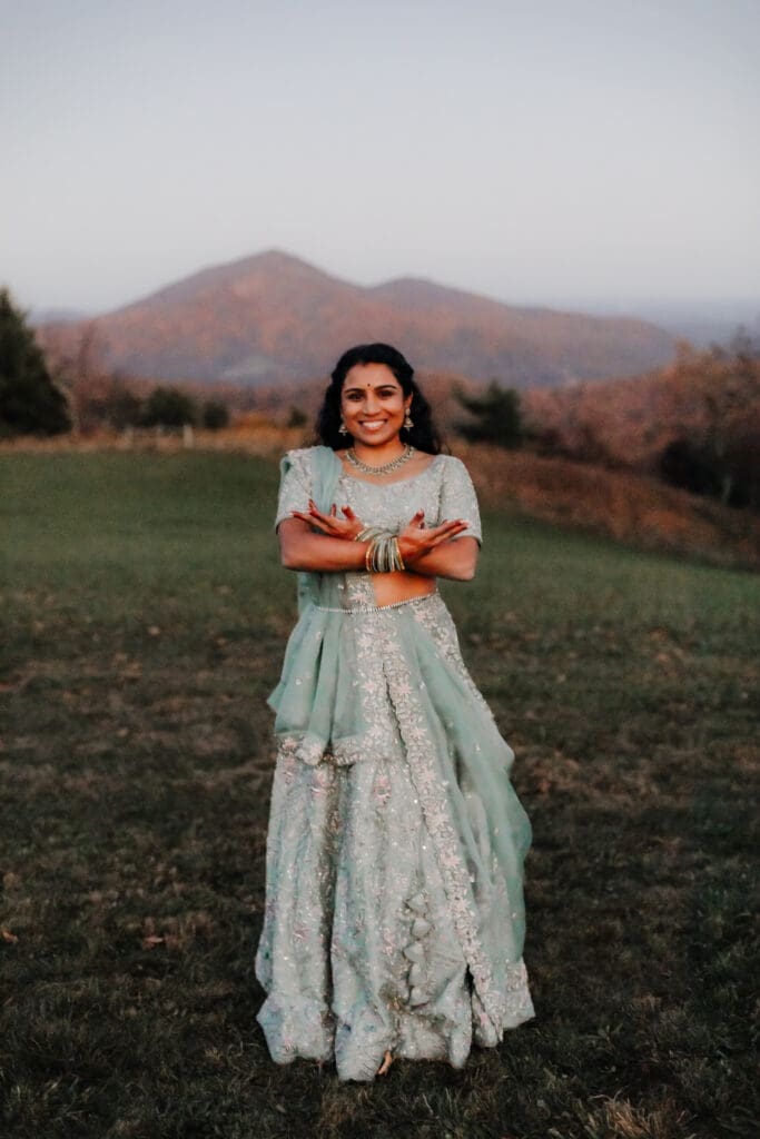 Indian bride in a Lehenga doing the Sangeet dance in green wedding attire in a field of green grass with a rust colored mountain in the background with blue rolling mountains in the background.