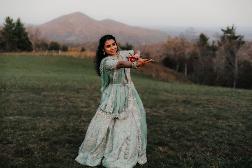 Indian bride in a Lehenga doing the Sangeet dance in green wedding attire in a field of green grass with a rust colored mountain in the background with blue rolling mountains in the background.
