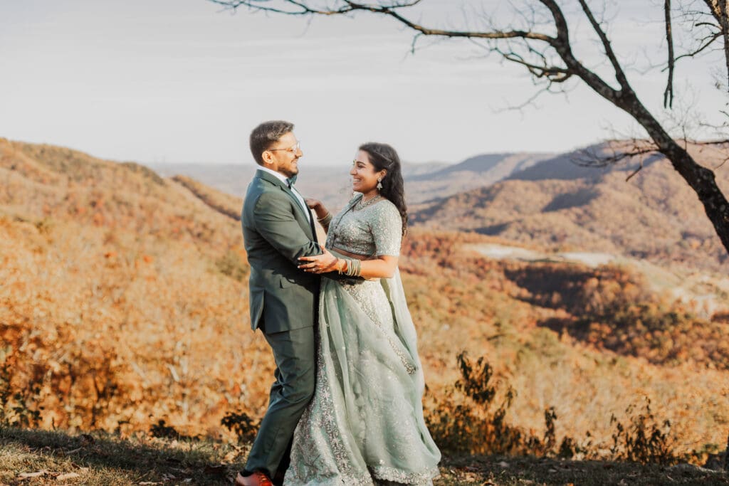 Indian couple in a in green wedding tux and lehenga in a field of green grass with a rust colored mountain in the background with blue rolling mountains in the background. They are standing close and kissing.