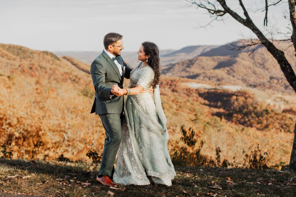 Indian couple in a in green wedding tux and lehenga in a field of green grass with a rust colored mountain in the background with blue rolling mountains in the background. They are standing close and kissing.