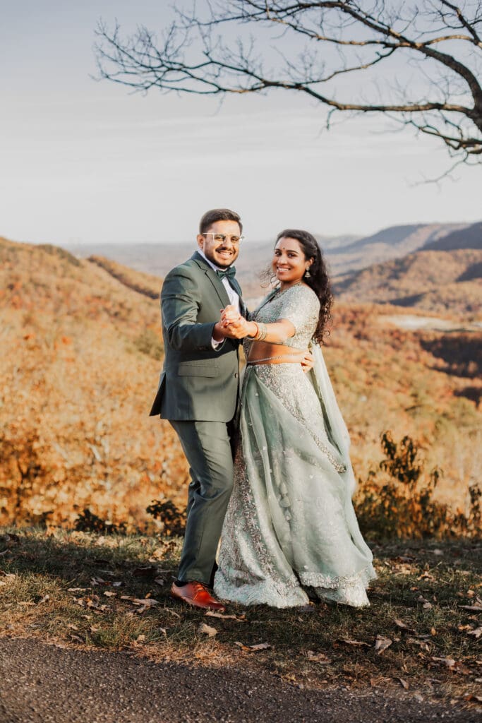 Indian couple in a in green wedding tux and lehenga in a field of green grass with a rust colored mountain in the background with blue rolling mountains in the background. They are standing close and kissing.