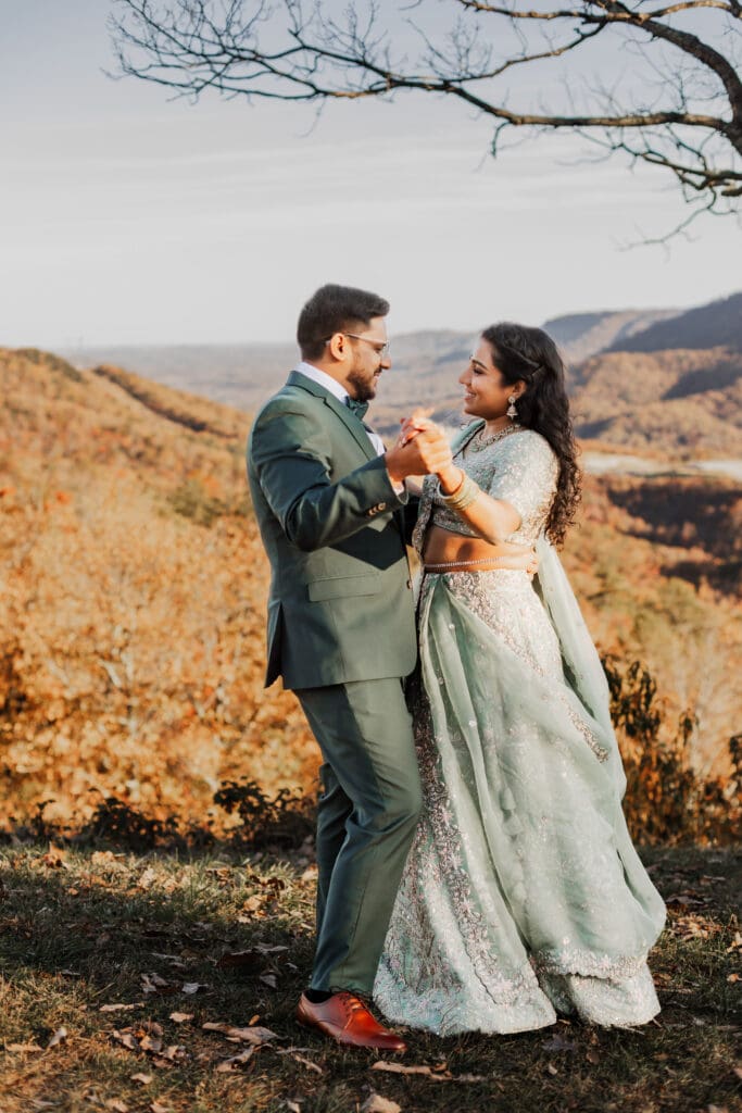 Indian couple in a in green wedding tux and lehenga in a field of green grass with a rust colored mountain in the background with blue rolling mountains in the background. They are standing close and kissing.