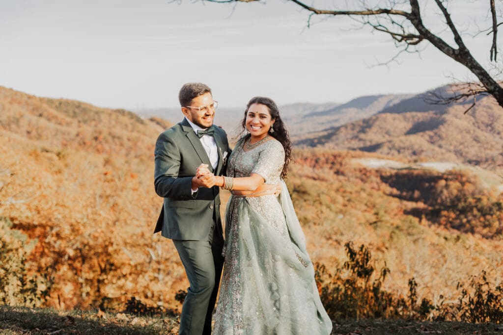 Indian couple in a in green wedding tux and lehenga in a field of green grass with a rust colored mountain in the background with blue rolling mountains in the background. They are standing close and kissing.