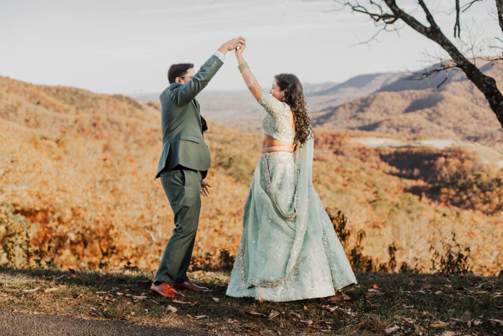 Indian couple in a in green wedding tux and lehenga in a field of green grass with a rust colored mountain in the background with blue rolling mountains in the background. They are standing close and kissing.