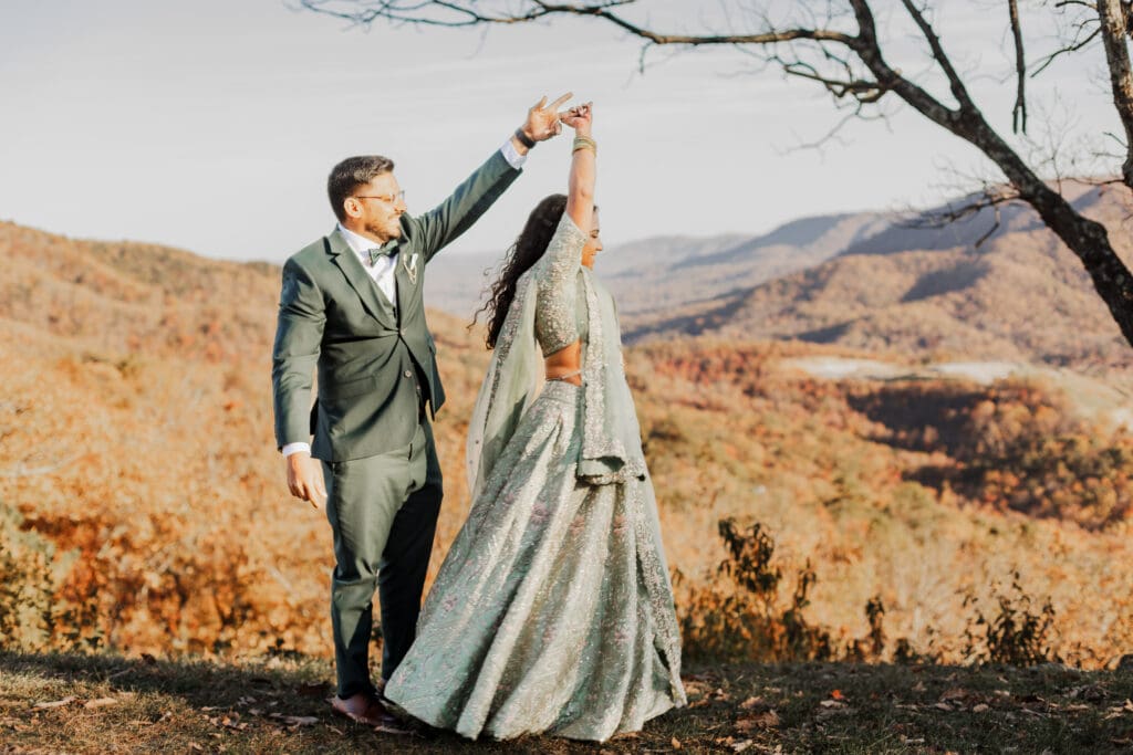 Indian couple in a in green wedding tux and lehenga in a field of green grass with a rust colored mountain in the background with blue rolling mountains in the background. They are standing close and kissing.