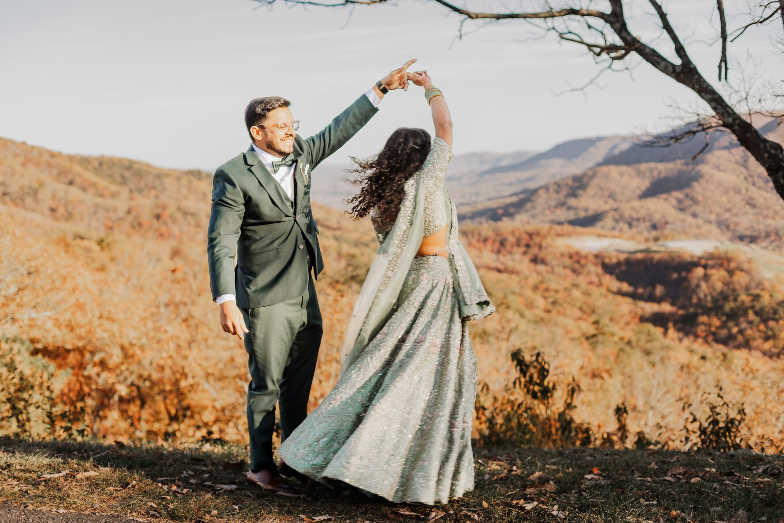 Indian couple in a in green wedding tux and lehenga in a field of green grass with a rust colored mountain in the background with blue rolling mountains in the background. They are standing close and kissing.