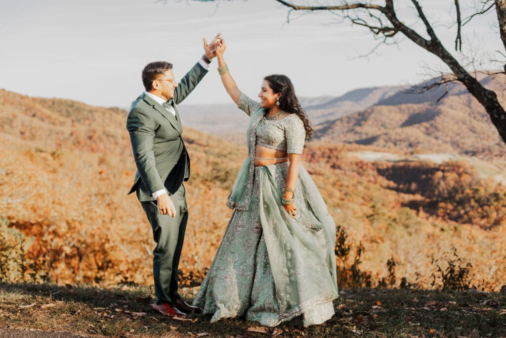 Indian couple in a in green wedding tux and lehenga in a field of green grass with a rust colored mountain in the background with blue rolling mountains in the background. They are standing close and kissing.