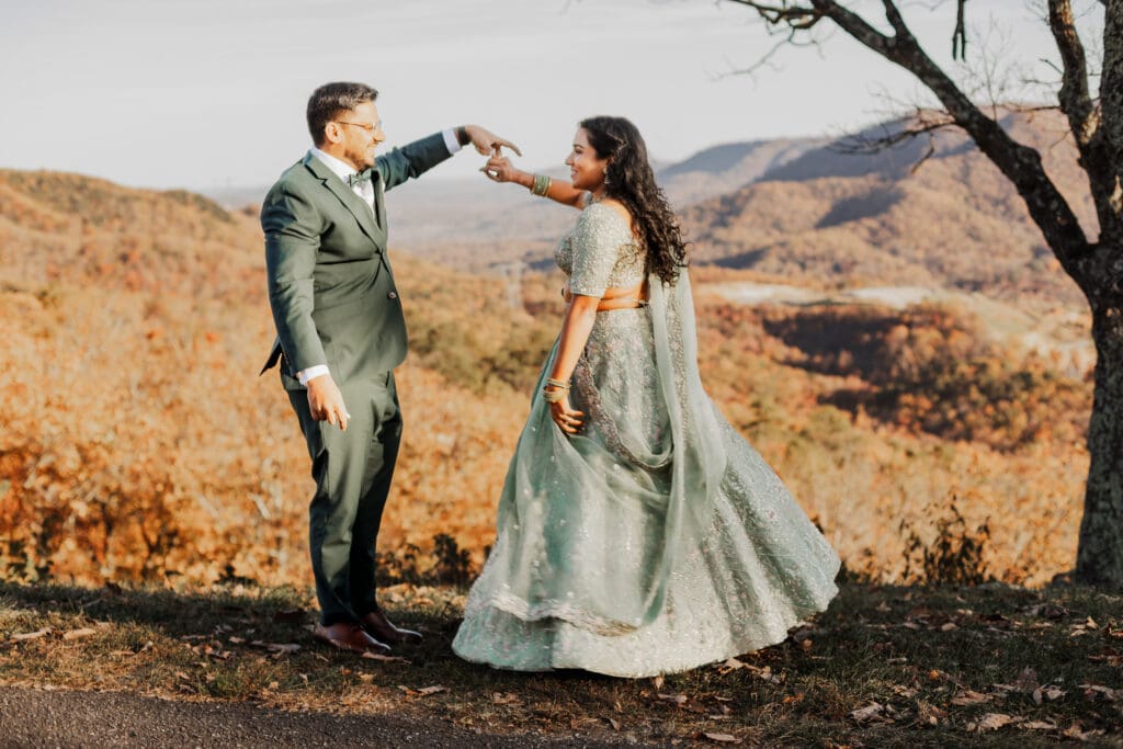 Indian couple in a in green wedding tux and lehenga in a field of green grass with a rust colored mountain in the background with blue rolling mountains in the background. They are standing close and kissing.