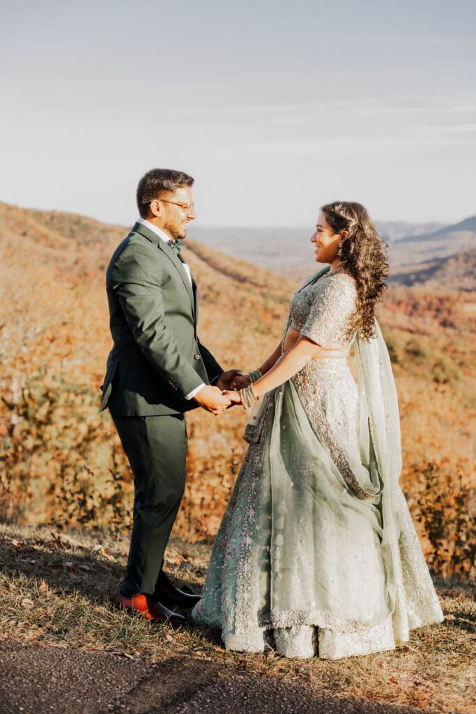 Indian couple in a in green wedding tux and lehenga in a field of green grass with a rust colored mountain in the background with blue rolling mountains in the background. They are standing close and kissing.