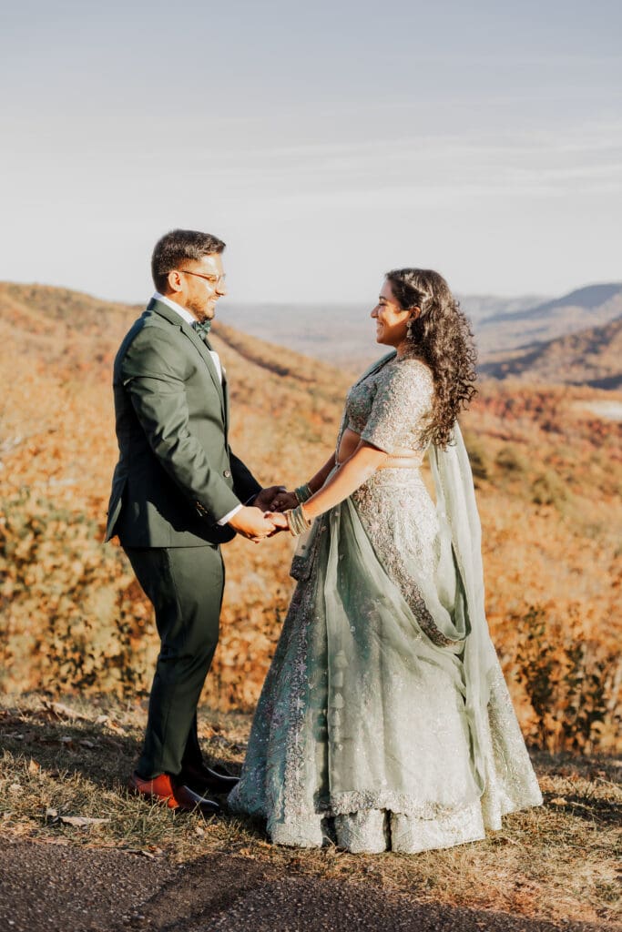 Indian couple in a in green wedding tux and lehenga in a field of green grass with a rust colored mountain in the background with blue rolling mountains in the background. They are standing close and kissing.