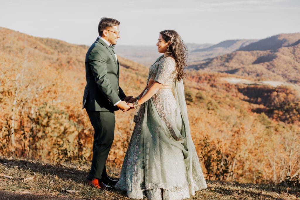 Indian couple in a in green wedding tux and lehenga in a field of green grass with a rust colored mountain in the background with blue rolling mountains in the background. They are standing close and kissing.
