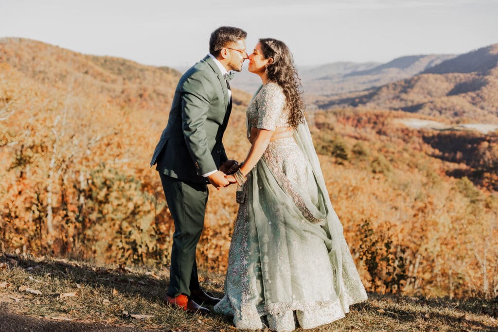 Indian couple in a in green wedding tux and lehenga in a field of green grass with a rust colored mountain in the background with blue rolling mountains in the background. They are standing close and kissing.
