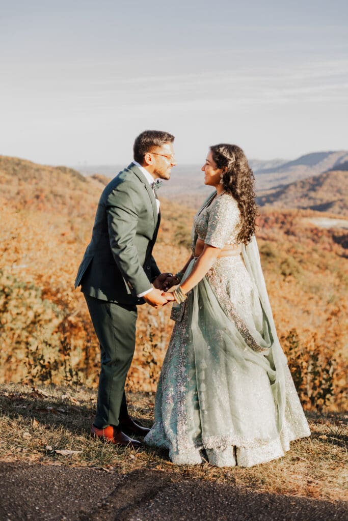 Indian couple in a in green wedding tux and lehenga in a field of green grass with a rust colored mountain in the background with blue rolling mountains in the background. They are standing close and kissing.