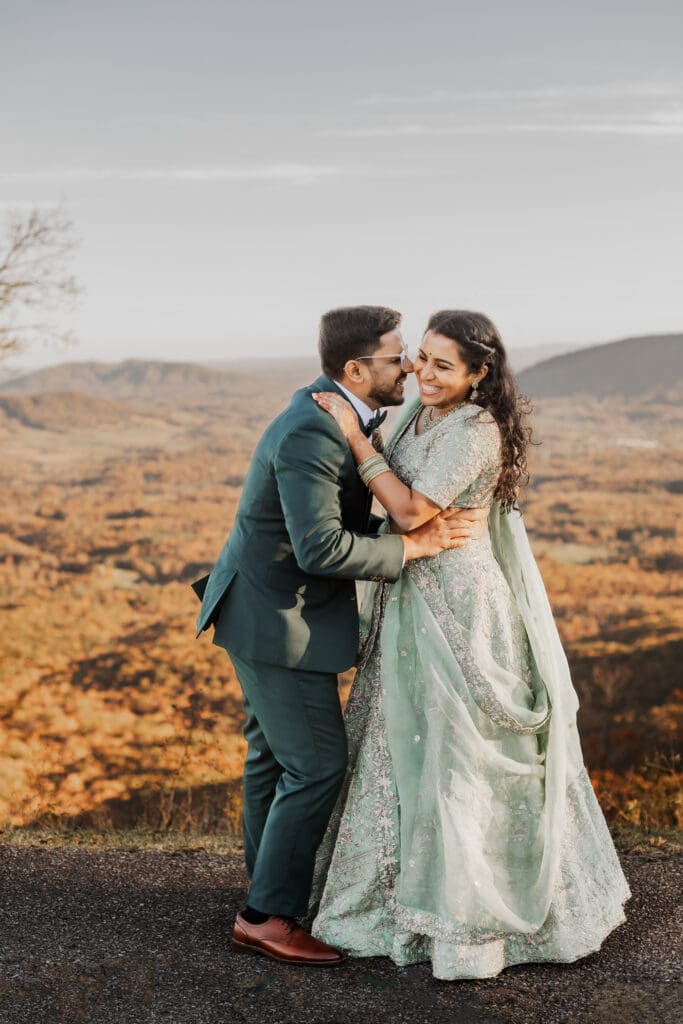 Indian couple in a in green wedding tux and lehenga in a field of green grass with a rust colored mountain in the background with blue rolling mountains in the background. They are standing close and kissing.