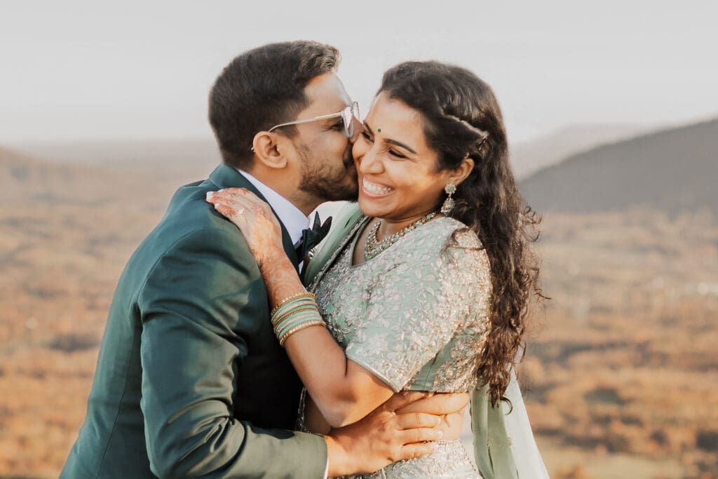 Indian couple in a in green wedding tux and lehenga in a field of green grass with a rust colored mountain in the background with blue rolling mountains in the background. They are standing close and kissing.