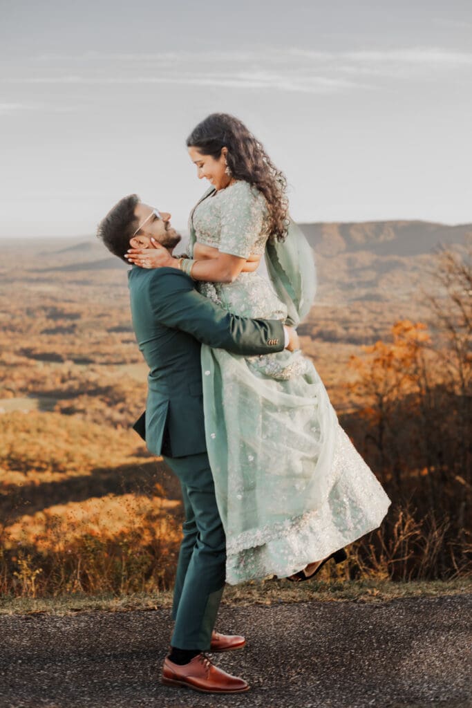 Indian couple in a in green wedding tux and lehenga in a field of green grass with a rust colored mountain in the background with blue rolling mountains in the background. They are standing close and kissing.