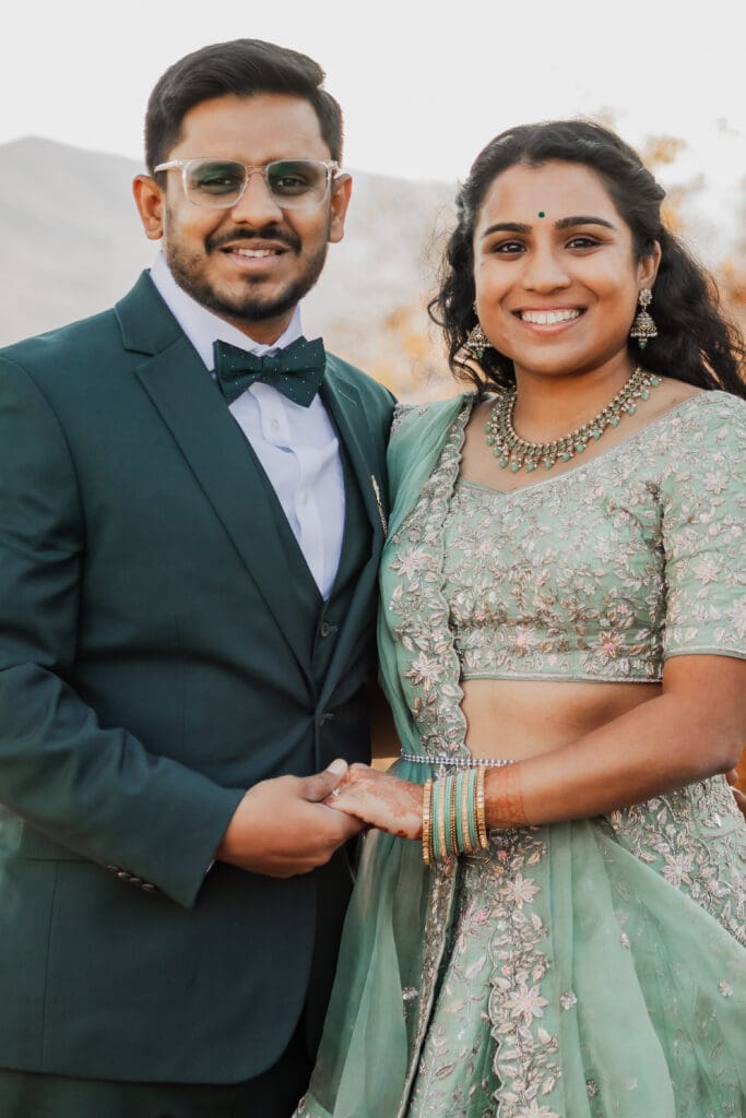 Indian couple in a in green wedding tux and lehenga in a field of green grass with a rust colored mountain in the background with blue rolling mountains in the background. They are standing close and kissing.