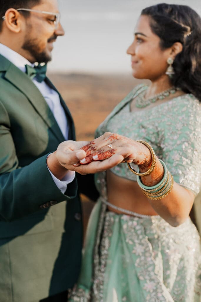 Indian couple in a in green wedding tux and lehenga in a field of green grass with a rust colored mountain in the background with blue rolling mountains in the background. They are standing close and kissing.