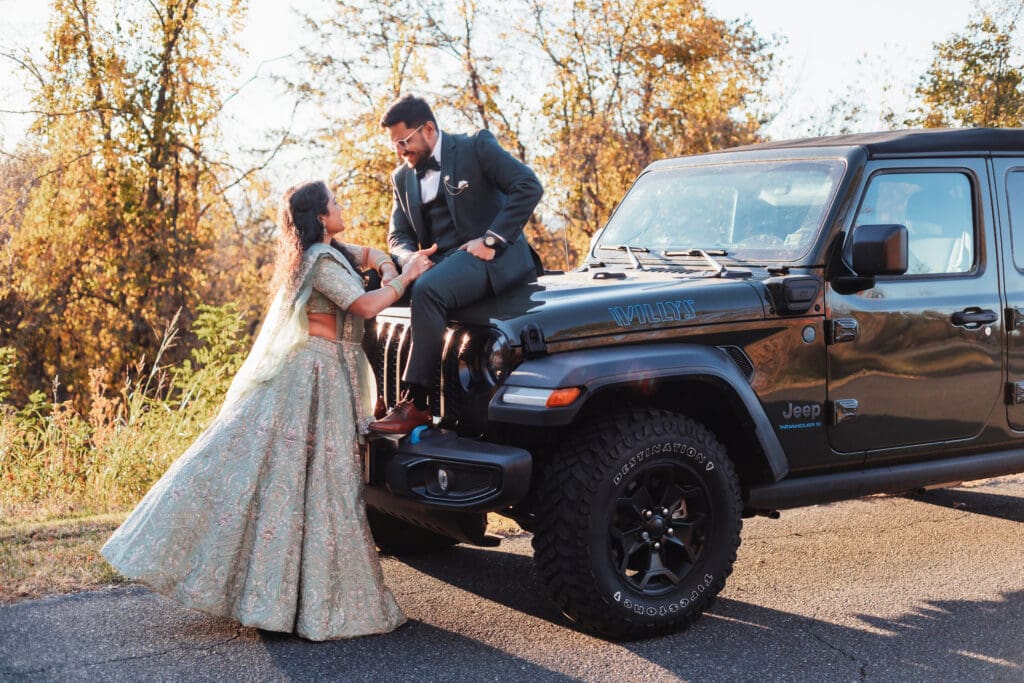 Indian couple in a in green wedding tux and lehenga in a field of green grass with a rust colored mountain in the background with blue rolling mountains in the background. They are standing close and kissing.
