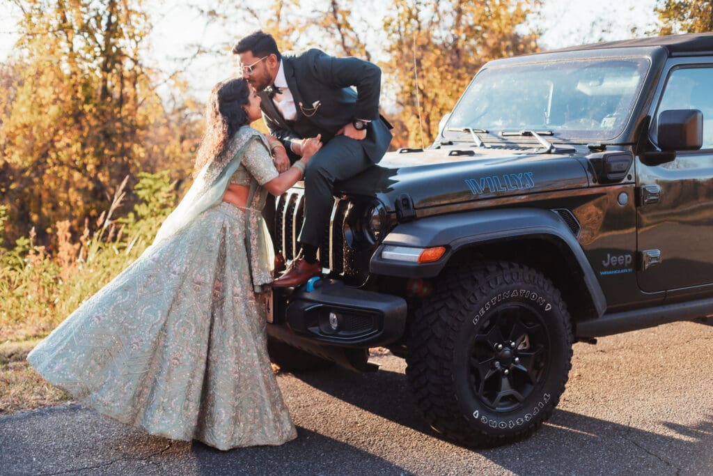 Indian couple in a in green wedding tux and lehenga in a field of green grass with a rust colored mountain in the background with blue rolling mountains in the background. They are standing close and kissing.