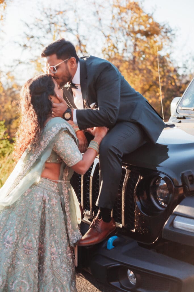 Indian couple in a in green wedding tux and lehenga in a field of green grass with a rust colored mountain in the background with blue rolling mountains in the background. They are standing close and kissing.