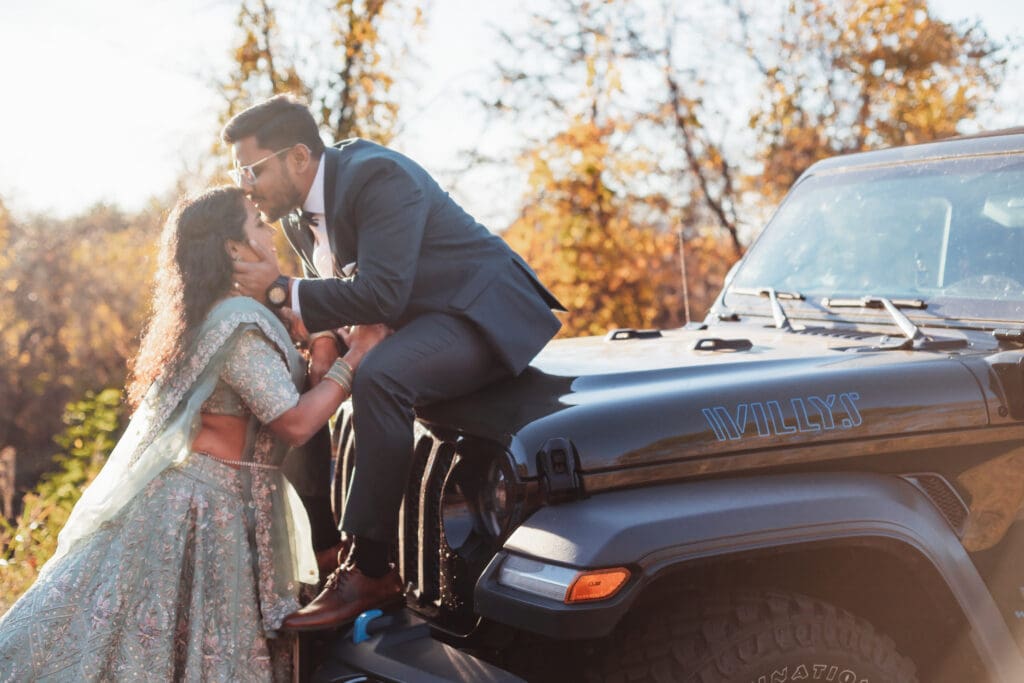 Indian couple in a in green wedding tux and lehenga in a field of green grass with a rust colored mountain in the background with blue rolling mountains in the background. They are standing close and kissing.