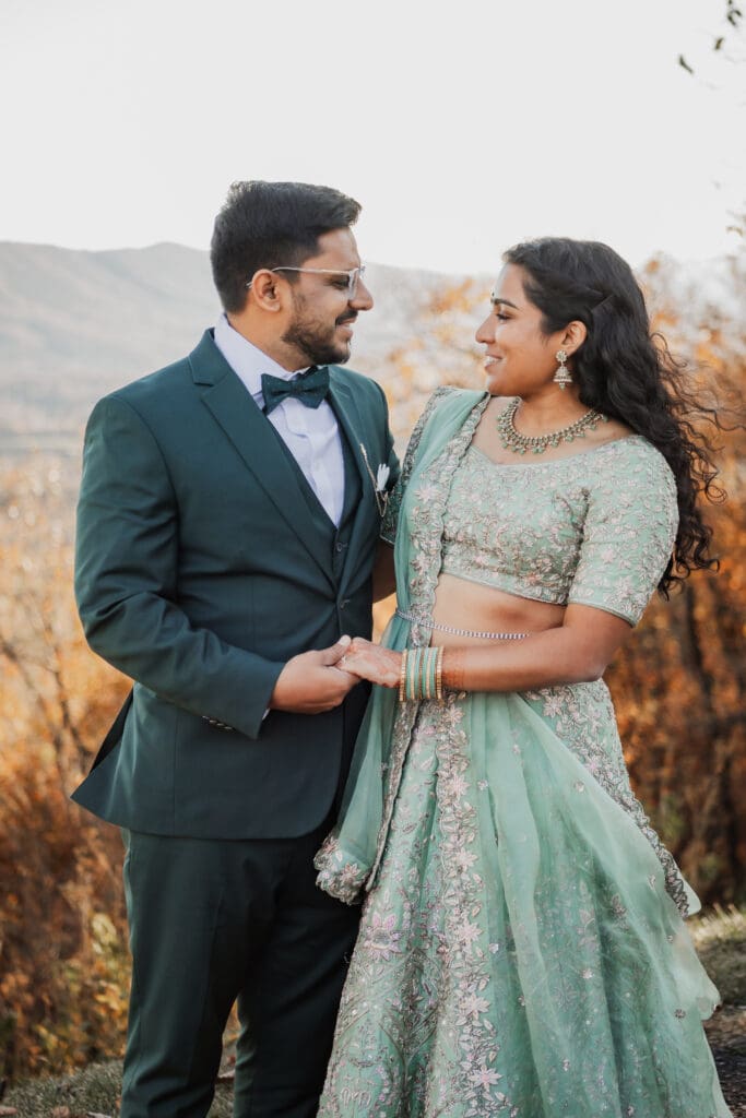 Indian couple in a in green wedding tux and lehenga in a field of green grass with a rust colored mountain in the background with blue rolling mountains in the background. They are standing close and kissing.
