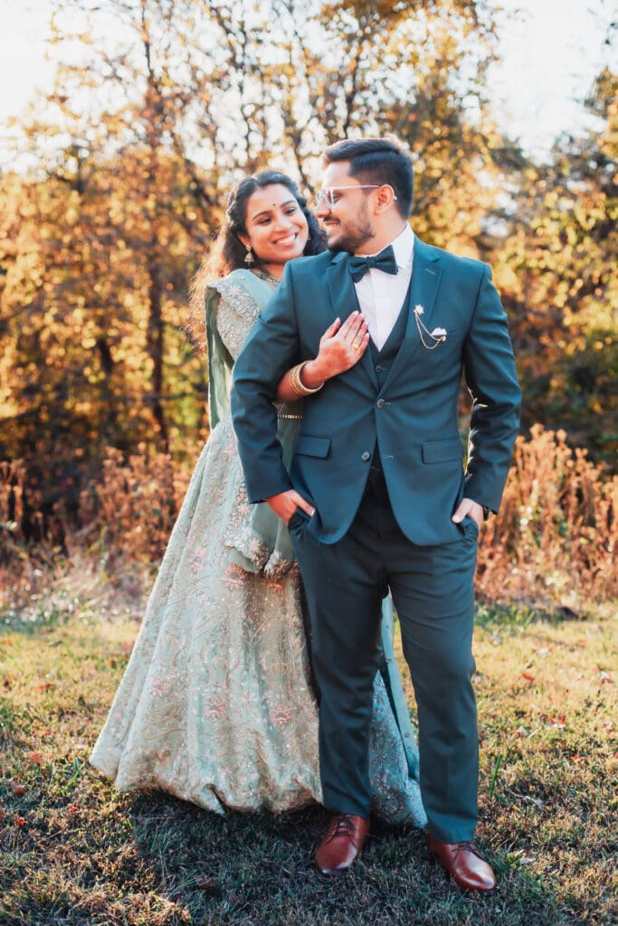 Indian couple in a in green wedding tux and lehenga in a field of green grass with a rust colored mountain in the background with blue rolling mountains in the background. They are standing close and kissing.