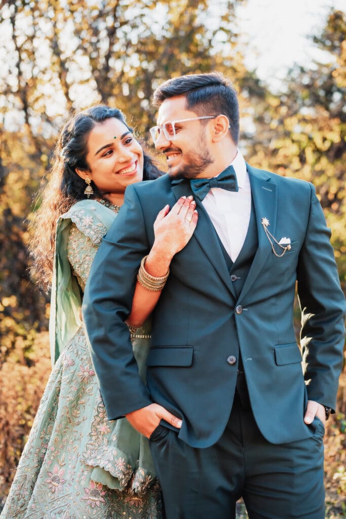Indian couple in a in green wedding tux and lehenga in a field of green grass with a rust colored mountain in the background with blue rolling mountains in the background. They are standing close and kissing.
