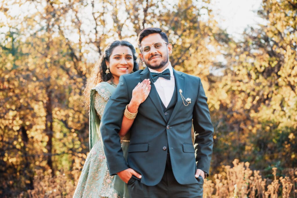 Indian couple in a in green wedding tux and lehenga in a field of green grass with a rust colored mountain in the background with blue rolling mountains in the background. They are standing close and kissing.