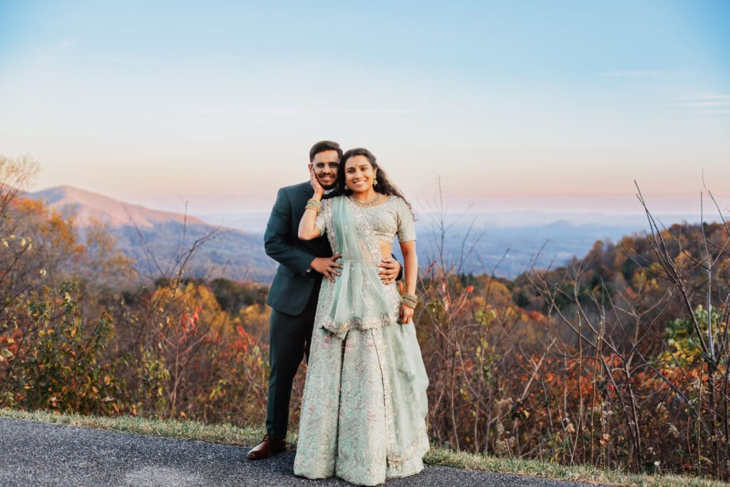 Indian couple in a in green wedding tux and lehenga in a field of green grass with a rust colored mountain in the background with blue rolling mountains in the background. They are standing close and kissing.