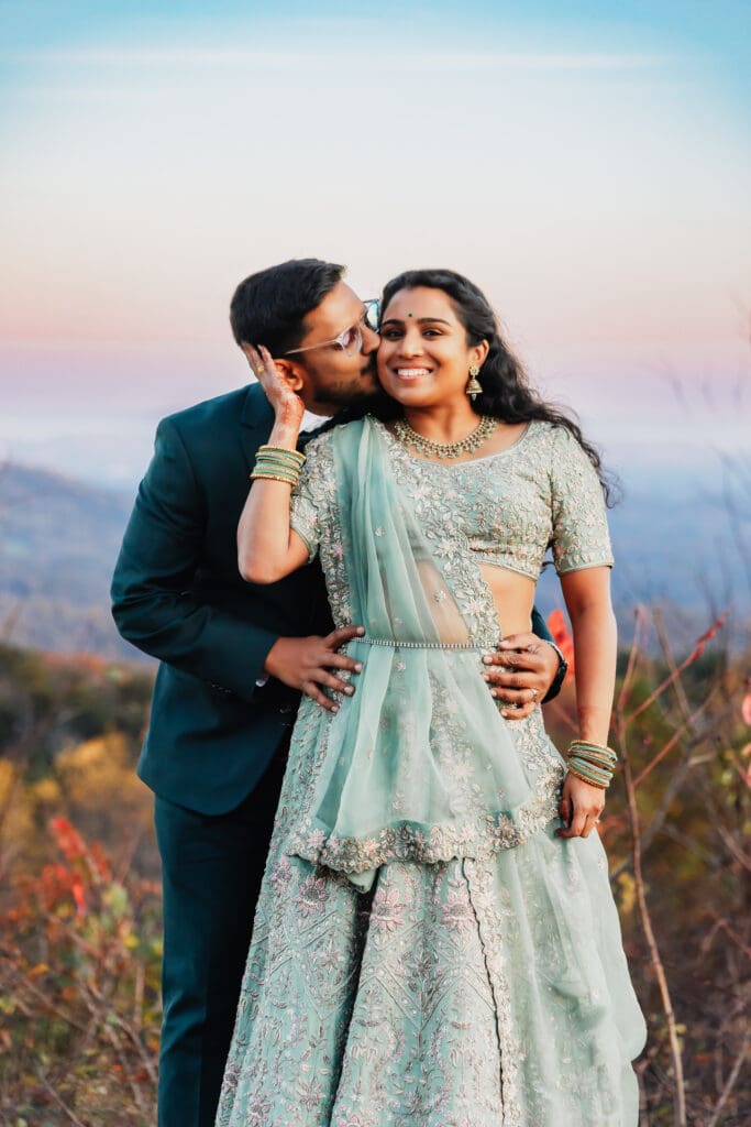 Indian couple in a in green wedding tux and lehenga in a field of green grass with a rust colored mountain in the background with blue rolling mountains in the background. They are standing close and kissing.