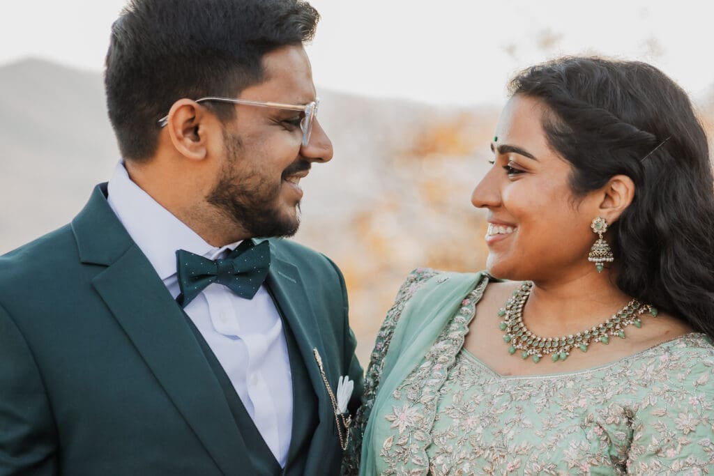Indian couple in a in green wedding tux and lehenga in a field of green grass with a rust colored mountain in the background with blue rolling mountains in the background. They are standing close and kissing.