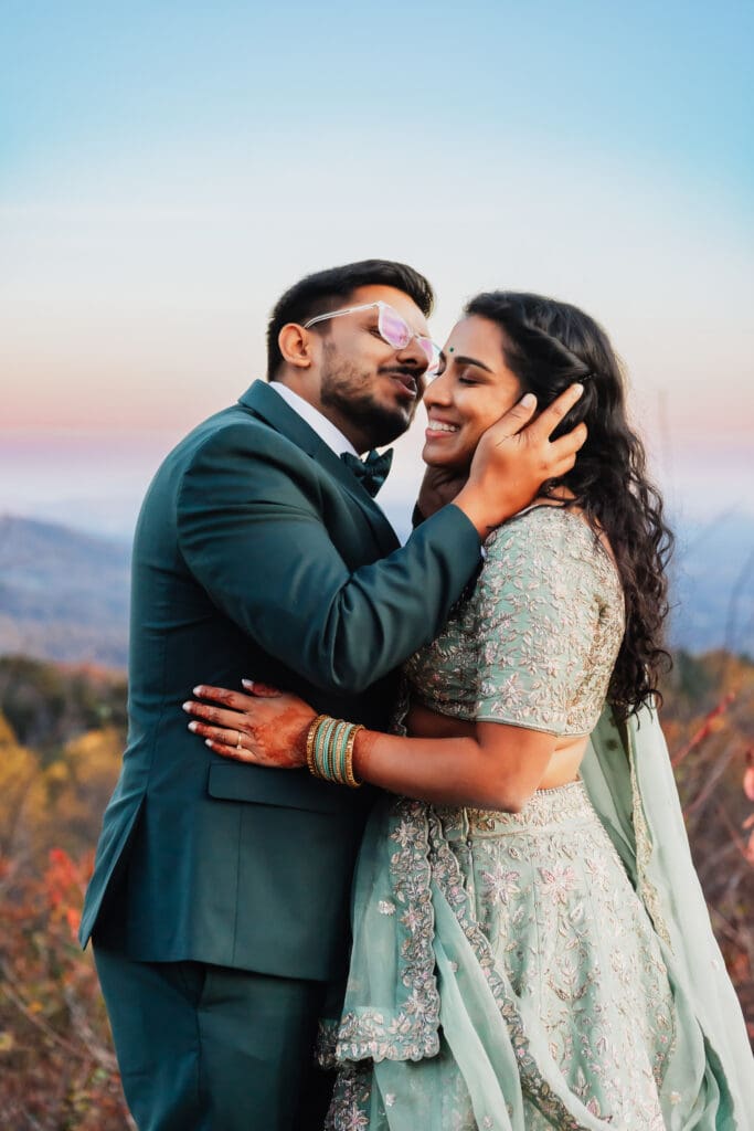 Indian couple in a in green wedding tux and lehenga in a field of green grass with a rust colored mountain in the background with blue rolling mountains in the background. They are standing close and kissing.