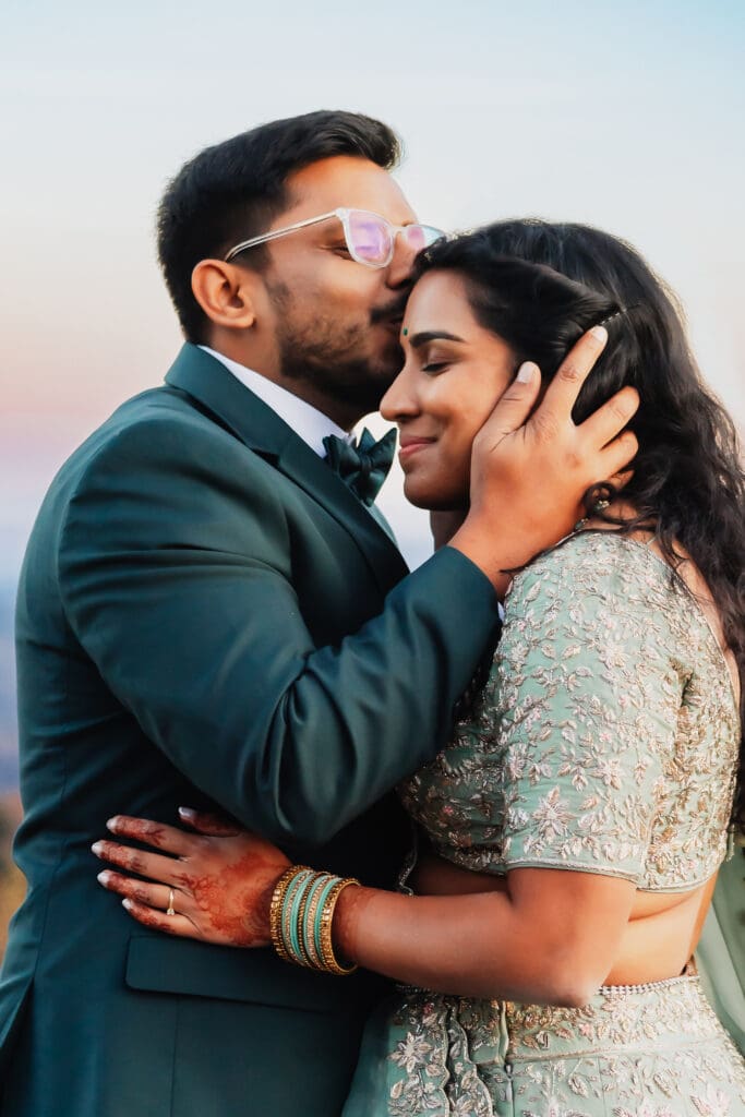Indian couple in a in green wedding tux and lehenga in a field of green grass with a rust colored mountain in the background with blue rolling mountains in the background. They are standing close and kissing.